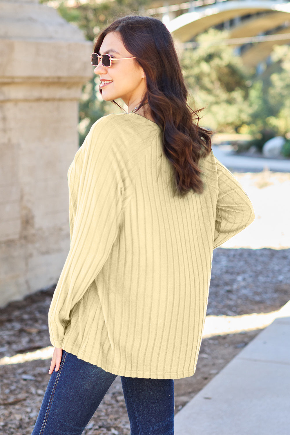 A woman with long brown hair wearing sunglasses and a Basic Bae full-size, slightly stretchy ribbed round neck long sleeve knit top in red, paired with blue jeans, is standing outdoors.