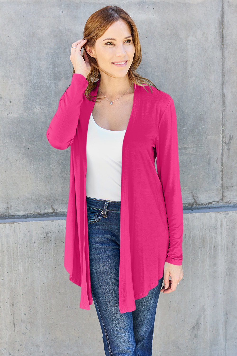 A woman wearing the Basic Bae Full Size Open Front Long Sleeve Cardigan in navy blue, paired with a white top and blue jeans, stands against a concrete wall. She touches her hair and gazes to the side, embodying a chic yet simple style. The slightly stretchy materials of the cardigan ensure comfort while maintaining its stylish simplicity. For easy care, machine wash it cold.