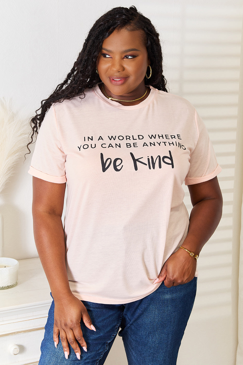 A person wears a Simply Love Slogan Graphic Cuffed T-Shirt in light pink, adorned with the inspiring message, "In a world where you can be anything, be kind," and featuring stylish cuffed sleeves.