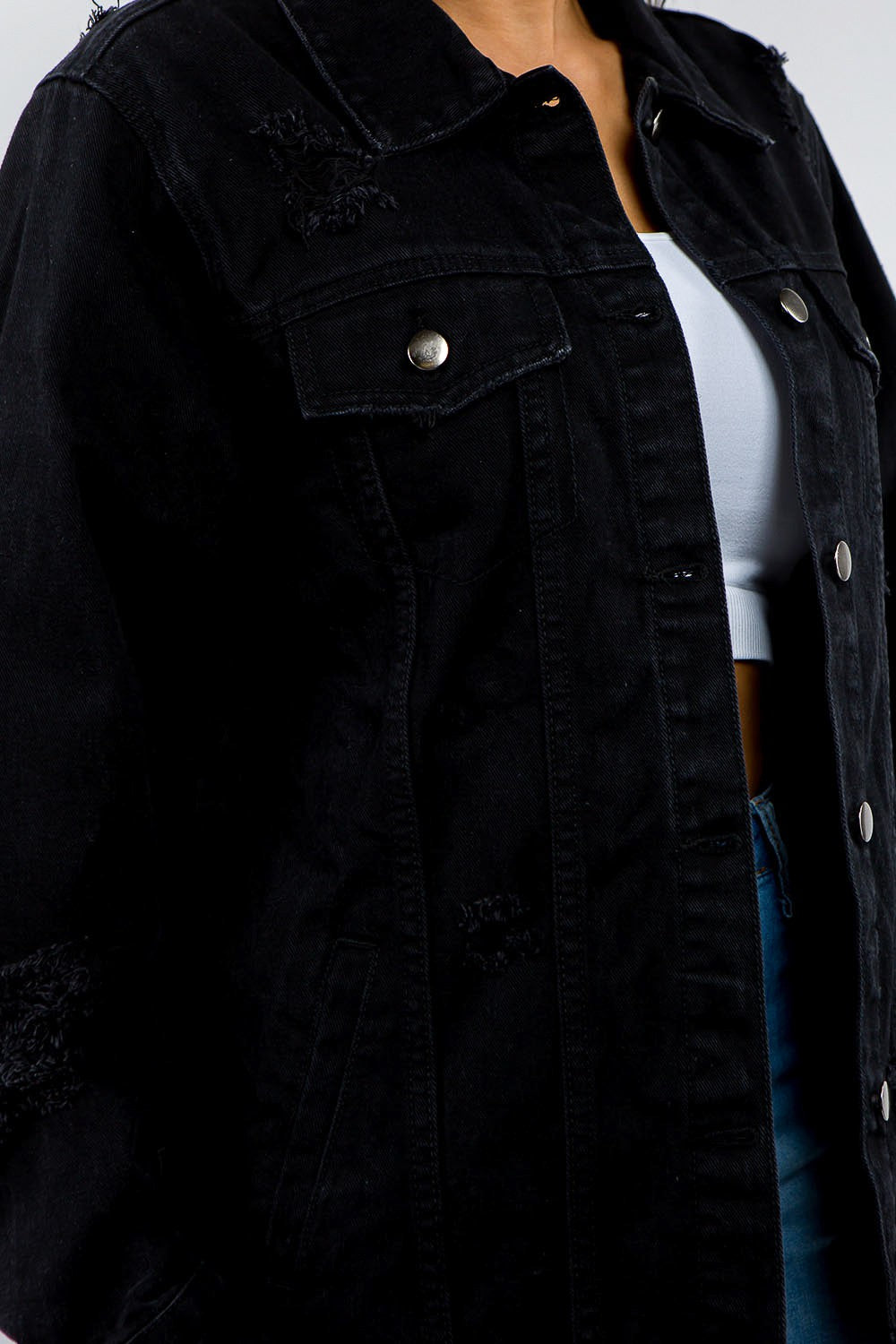 A woman stands against a plain background, epitomizing casual outfits with a touch of vintage look, wearing the American Bazi Full Size Button Up Distressed Denim Jacket over a white crop top and jeans.