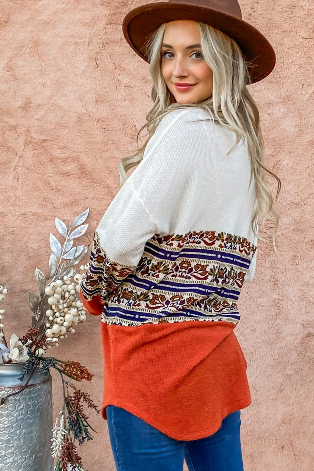 A woman with long blonde hair wearing a brown hat and the And The Why Ethnic Print Color Block Top stands against a peach wall next to a floral arrangement.