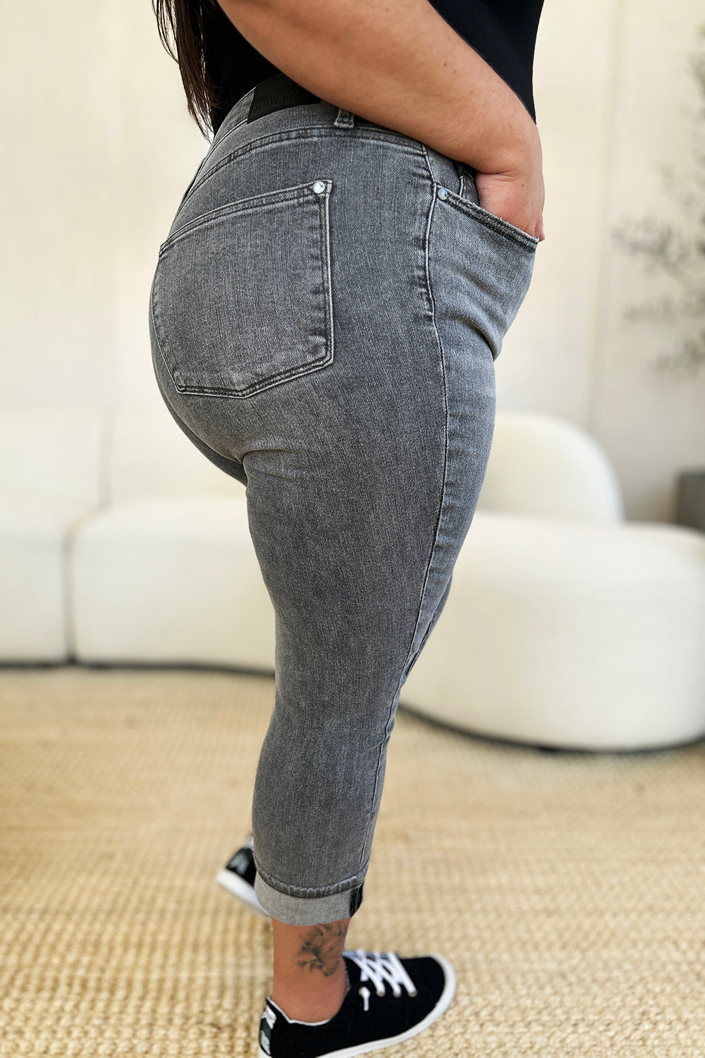 A person wearing Judy Blue Full Size Button Fly High Waist Cuffed Capris in gray, paired with a white top and white sneakers, stands on a beige carpeted floor.