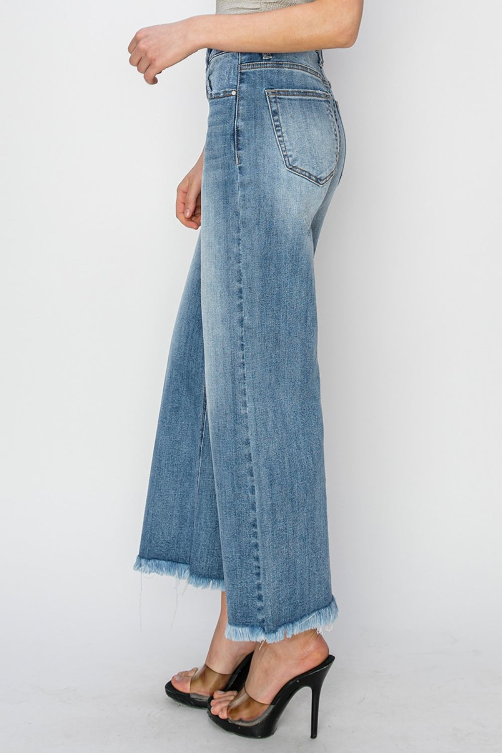 Person wearing RISEN Raw Hem Cropped Wide Leg Jeans in blue with a beige crop top. Only the lower half of the body is visible, paired with strappy black heels.