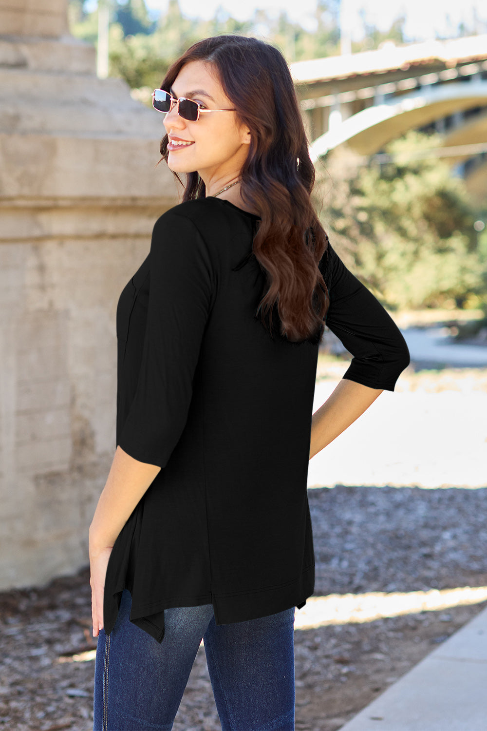 A woman stands outdoors wearing sunglasses, a Basic Bae Full Size Round Neck Pocketed T-Shirt with an asymmetrical hem and chest pocket made of rayon spandex, and blue jeans. She is smiling and has one hand in her pocket. Her basic style radiates effortless charm.