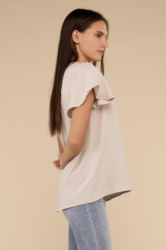 A woman with long straight hair models the Woven Airflow Flutter Sleeve Top in green, matched with light blue jeans, as she poses with her hands in her pockets against a plain beige background.