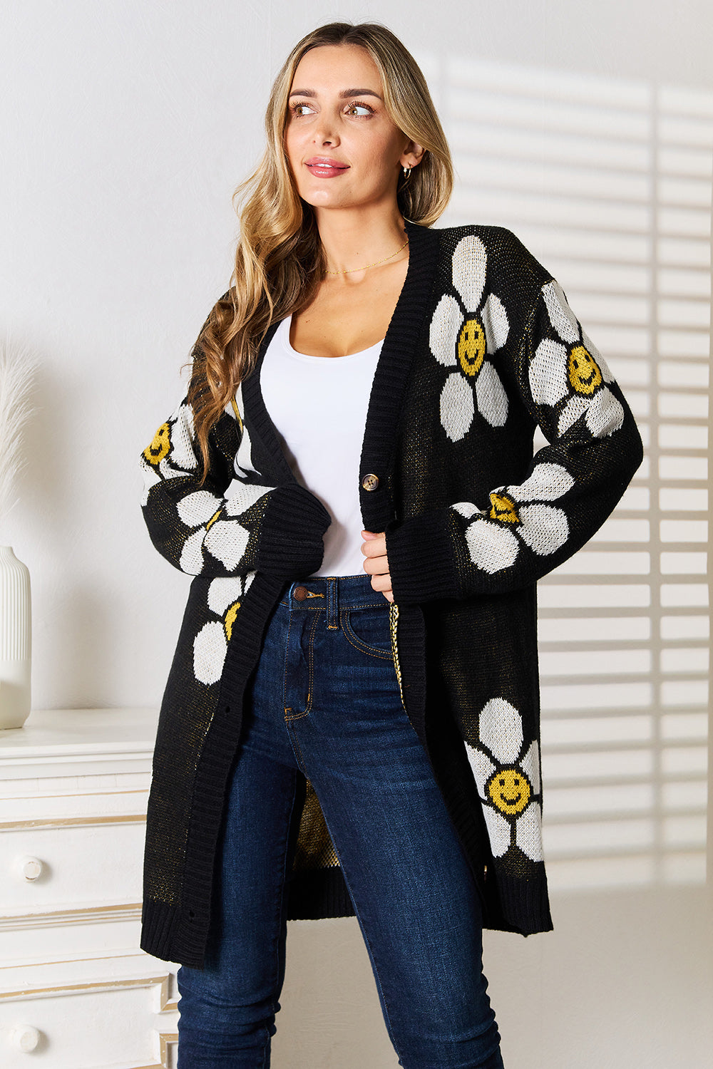 A woman stands indoors wearing the Perfee Floral Button Down Longline Cardigan, which features stylish black fabric adorned with white and yellow smiley face flowers. She pairs it with a white top and blue jeans, looking off to the side while smiling.