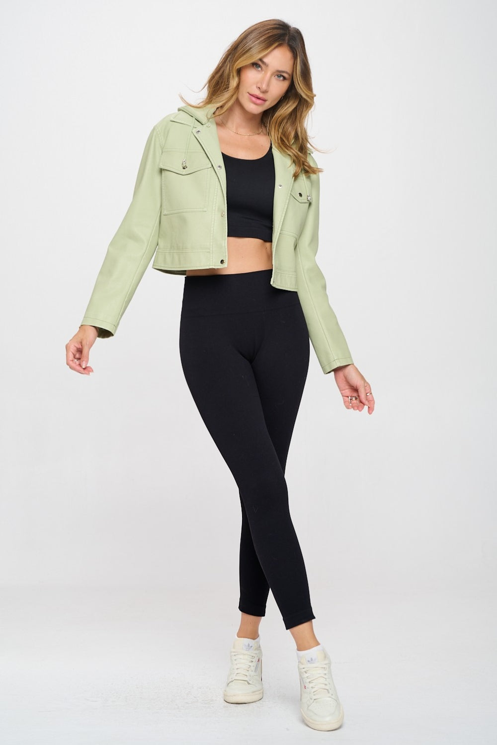 A person showcases a trendy Coalition LA Snap Down Cropped Hooded Jacket in light green over a sleek black outfit against a plain background.