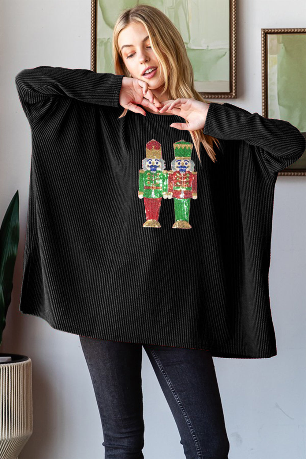 A woman wearing a black Heimish Sequin Nutcracker Long Sleeve Ribbed Top adorned with colorful figures on the front is standing indoors.