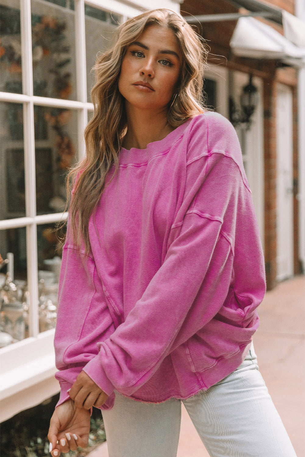 A woman stands with her back to the camera, wearing a long-sleeved Rose Exposed Seam Twist Open Back Oversized Sweatshirt and light blue jeans, showcasing a relaxed fit.