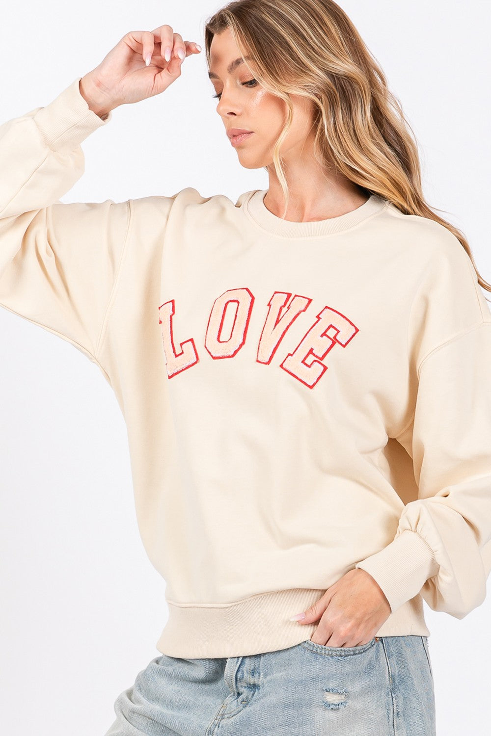 A woman dressed in a SAGE + FIG LOVE Path Applique Drop Shoulder Sweatshirt and a wide-brimmed hat is touching the brim of her hat with both hands, looking to the side and flaunting her cozy and stylish sweatshirt.