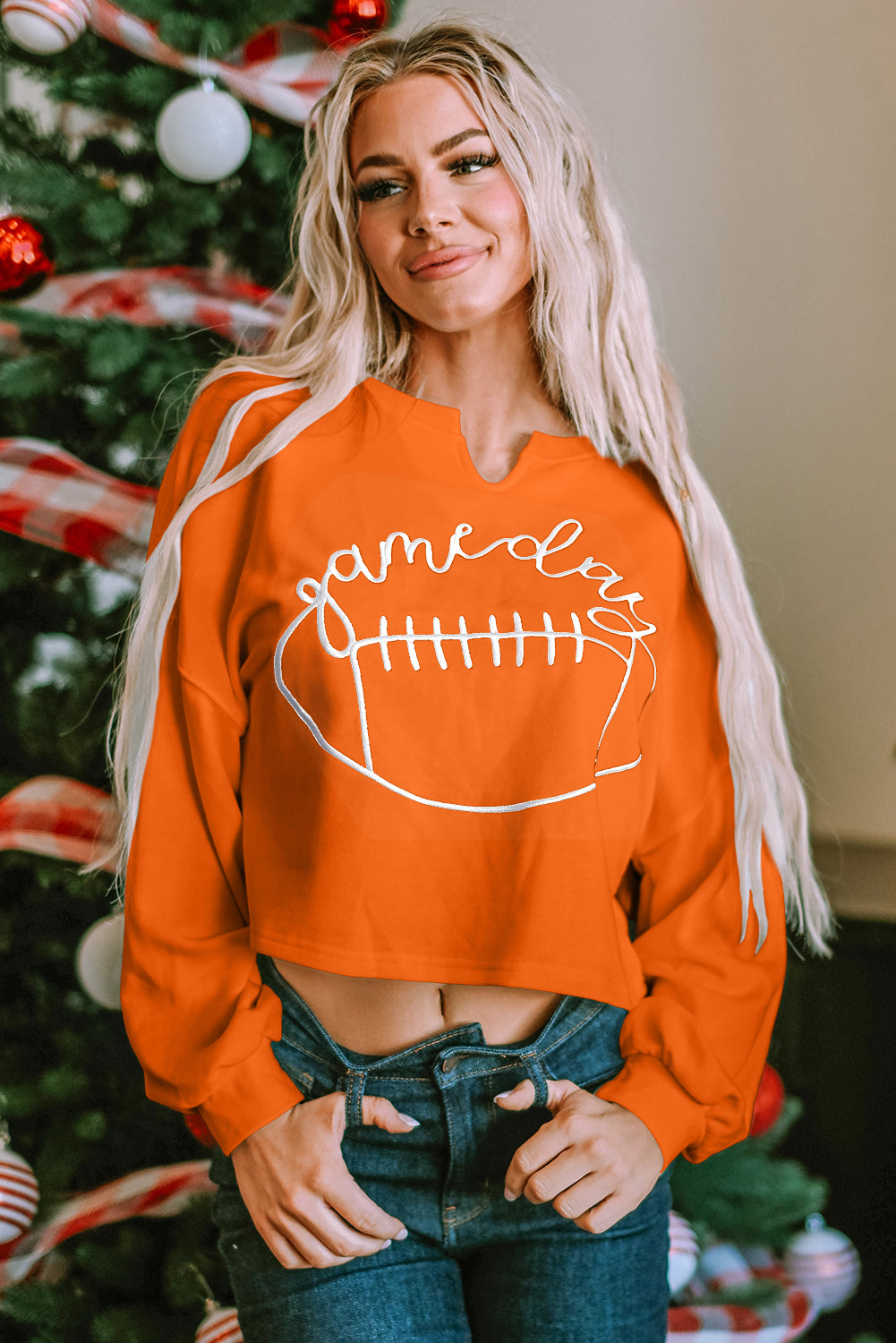 A woman with blonde hair, wearing an Orange Game Day Lettering Rugby Football Notched Neck Sweatshirt and blue jeans, stands next to a decorated Christmas tree.