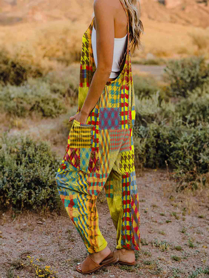 A woman stands outdoors in front of a rocky landscape, wearing the Full Size Printed V-Neck Sleeveless Jumpsuit and a brown brimmed hat. She is smiling and holding the brim of her hat with one hand, exuding the carefree vibe of this vibrant and colorful jumpsuit.