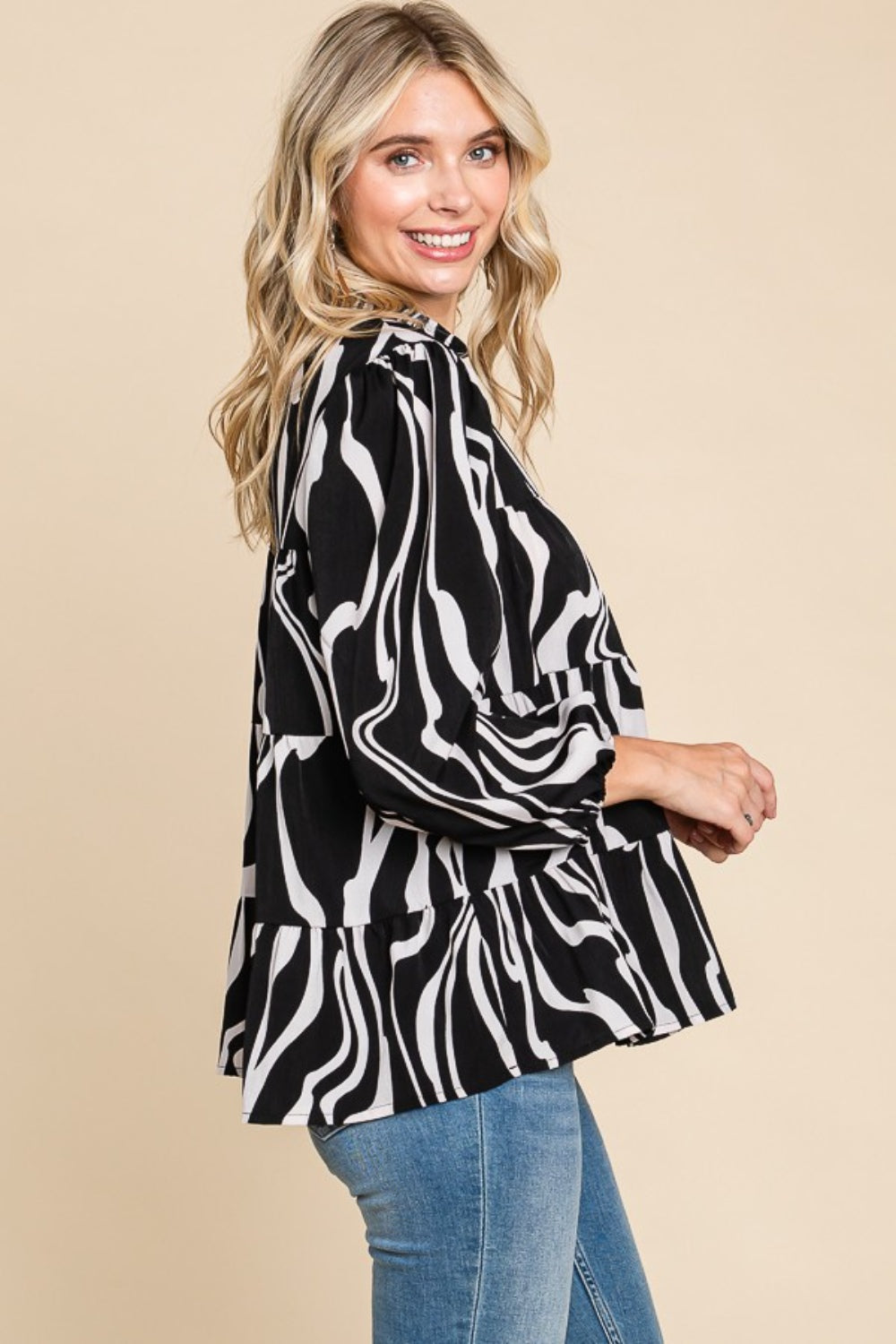 A person wearing a Culture Code Printed Smock Neck Tiered Blouse, featuring a black and white patterned design and long sleeves, paired with blue jeans, stands against a beige background.