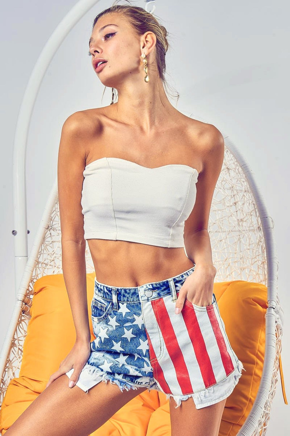 A person wearing BiBi US Flag Theme Bleached Denim Shorts and a white crop top stands with hands in pockets against a yellow background.