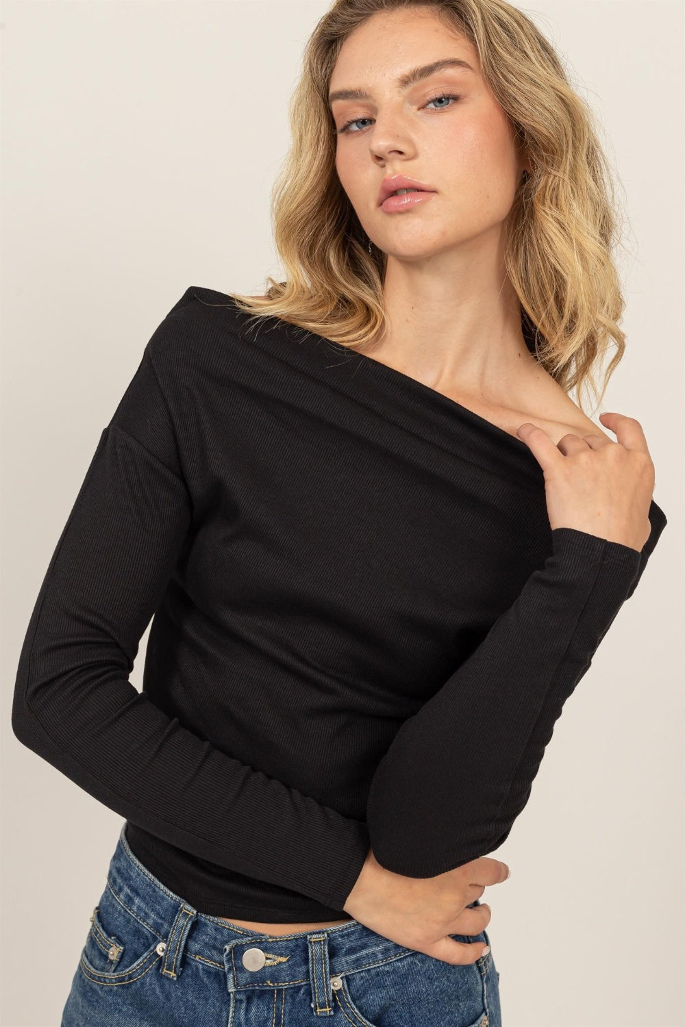 A person is posing gracefully, adorned in the HYFVE Off Shoulder Ribbed Knit Top, paired with blue jeans. Their hand rests elegantly on their neck against a neutral backdrop, highlighting a versatile wardrobe choice perfect for any occasion.