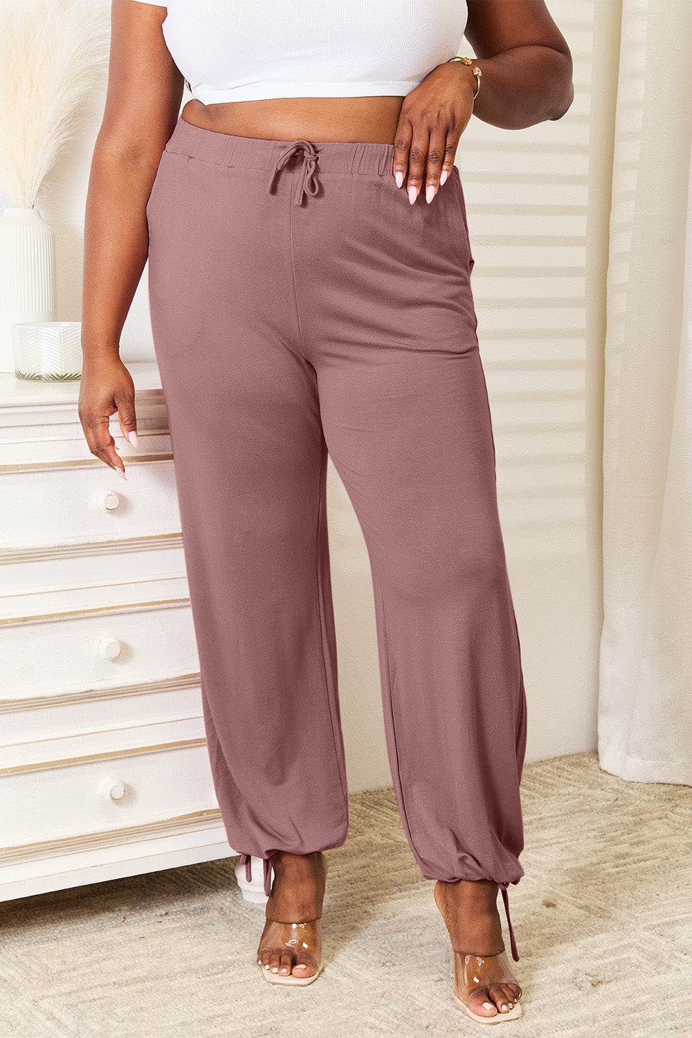 Person wearing Basic Bae Full Size Soft Rayon Drawstring Waist Pants with Pockets in teal, standing next to a white dresser with hands in pockets. These comfortable and stylish pants represent versatile everyday wear.