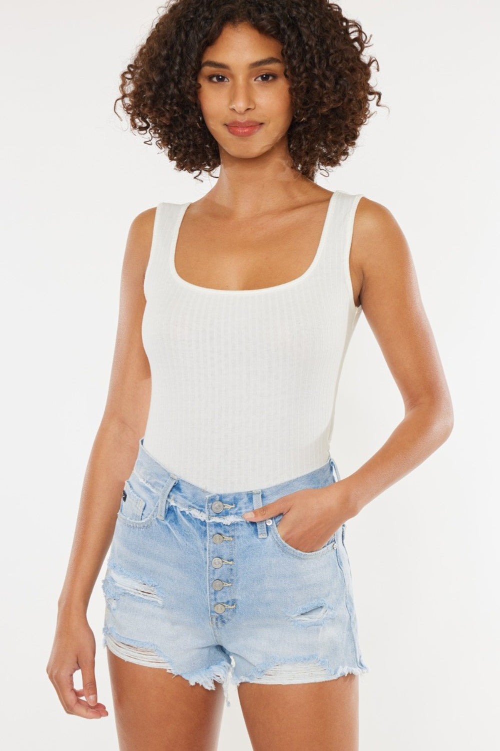 A person wearing a white tank top and Kancan Distressed Button Fly Denim Shorts poses against a plain background, showcasing a quintessential summer wardrobe staple.