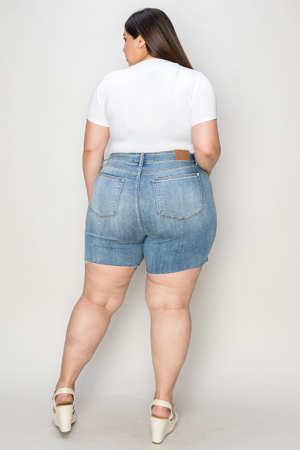 Side view of a person wearing a white shirt and Judy Blue Full Size High Waist Raw Hem Denim Shorts, standing indoors near a wooden cabinet. Summer fashion vibes radiate through the casual ensemble.