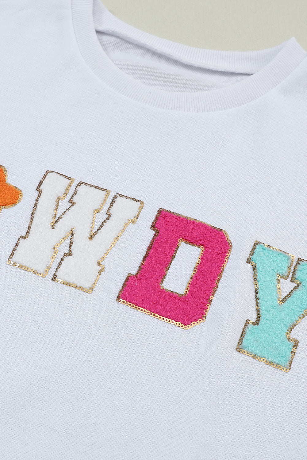 A person poses in the White Howdy Glitter Chenille Patch Graphic Casual Sweatshirt, adorned with colorful star patterns and the letters "WD." She wears light denim shorts and has shoulder-length blonde hair.