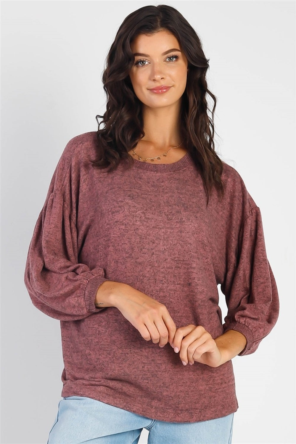 A person with long dark hair stands against a white background, smiling and raising one hand while wearing the trendy and stylish Cherish Apparel Drop Shoulder Puff Sleeve Top in burgundy paired with light blue jeans.