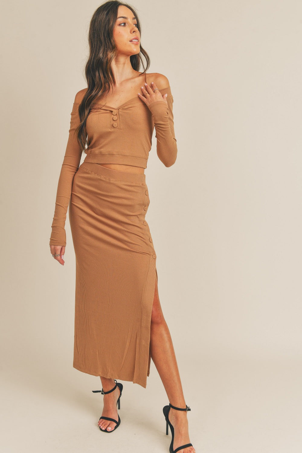 A woman stands against a plain background wearing the MABLE Button Detail Crop Top and Slit Midi Skirt Set, showcasing a camel-colored ensemble with off-shoulder, button details, and a side slit. She completes her look with black high-heeled sandals.