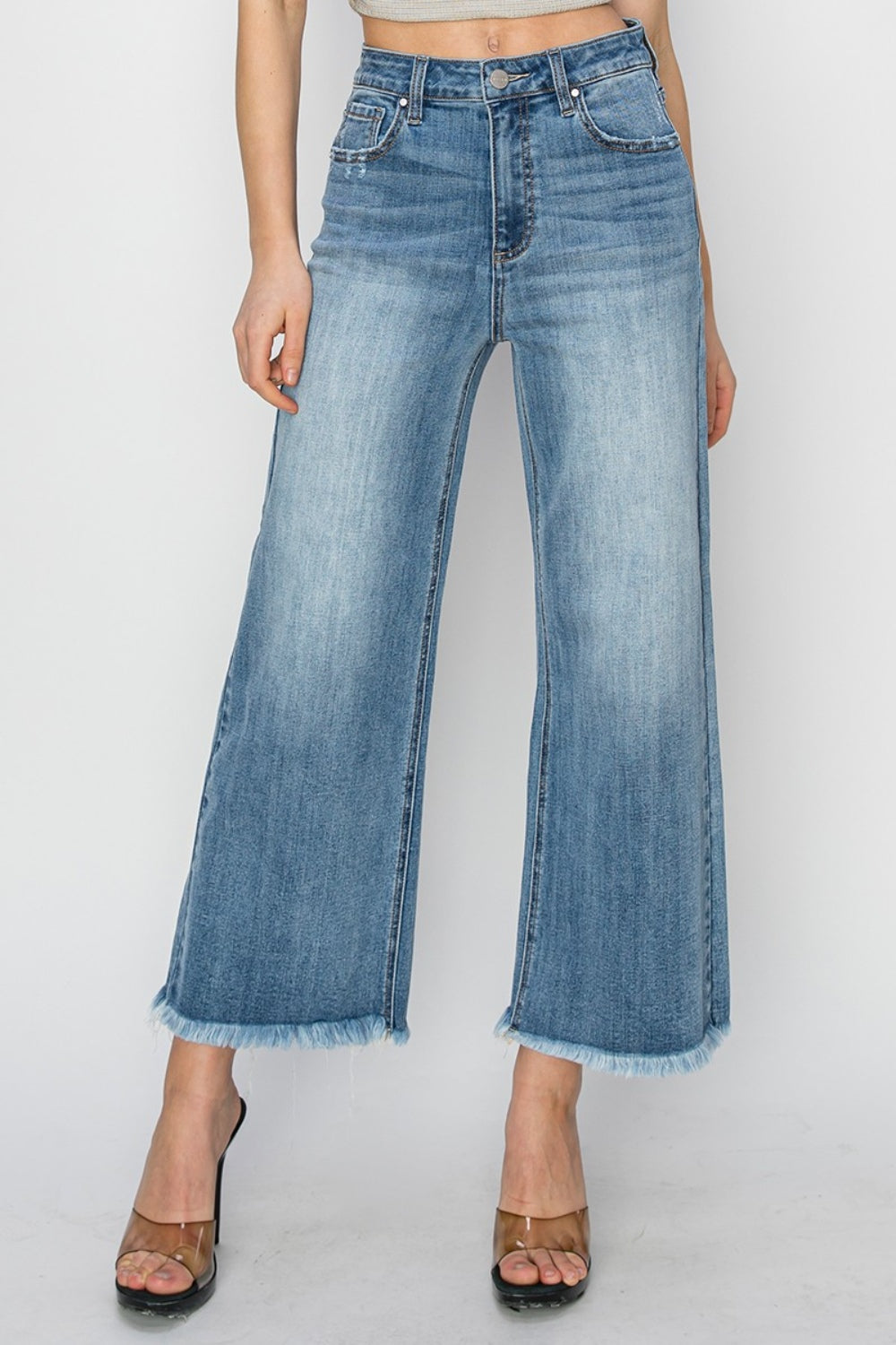 Person wearing RISEN Raw Hem Cropped Wide Leg Jeans in blue with a beige crop top. Only the lower half of the body is visible, paired with strappy black heels.