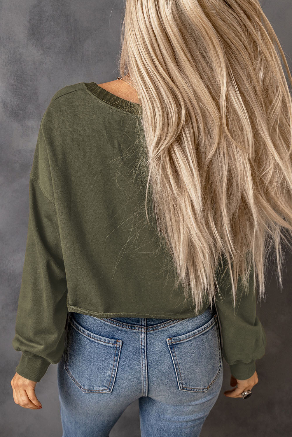 A person with long blonde hair stands with their back to the camera, wearing a faddish and cozy Green Drop Shoulder Cropped Sweatshirt and blue jeans.