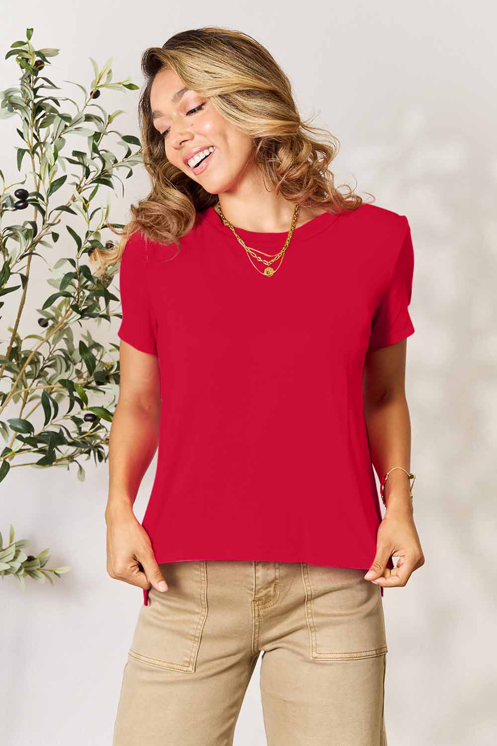 A woman stands next to an indoor plant, smiling, with one hand in her khaki pants pocket. In her pink Basic Bae Full Size Round Neck Short Sleeve T-Shirt and gold jewelry, she radiates casual elegance.