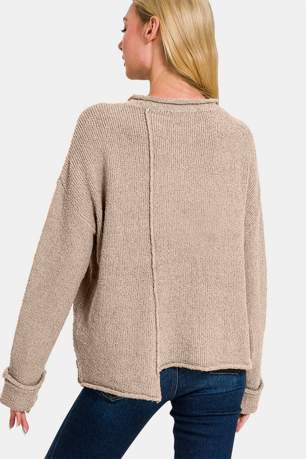 A woman with long blonde hair wears a trendy Zenana Asymmetric Hem Drop Shoulder Sweater in beige and blue jeans, posing with one hand touching her chin and the other holding the hem of her sweater.