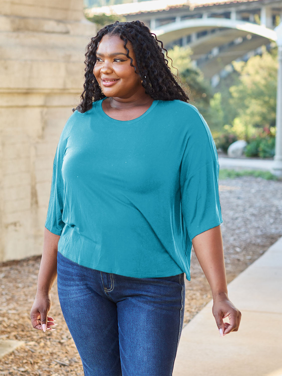 A person with long dark hair and a bright smile stands outdoors, showcasing their basic style in a loose, slightly stretchy *Basic Bae Full Size Round Neck Drop Shoulder T-Shirt* in pink and blue jeans, with one hand on their hip.