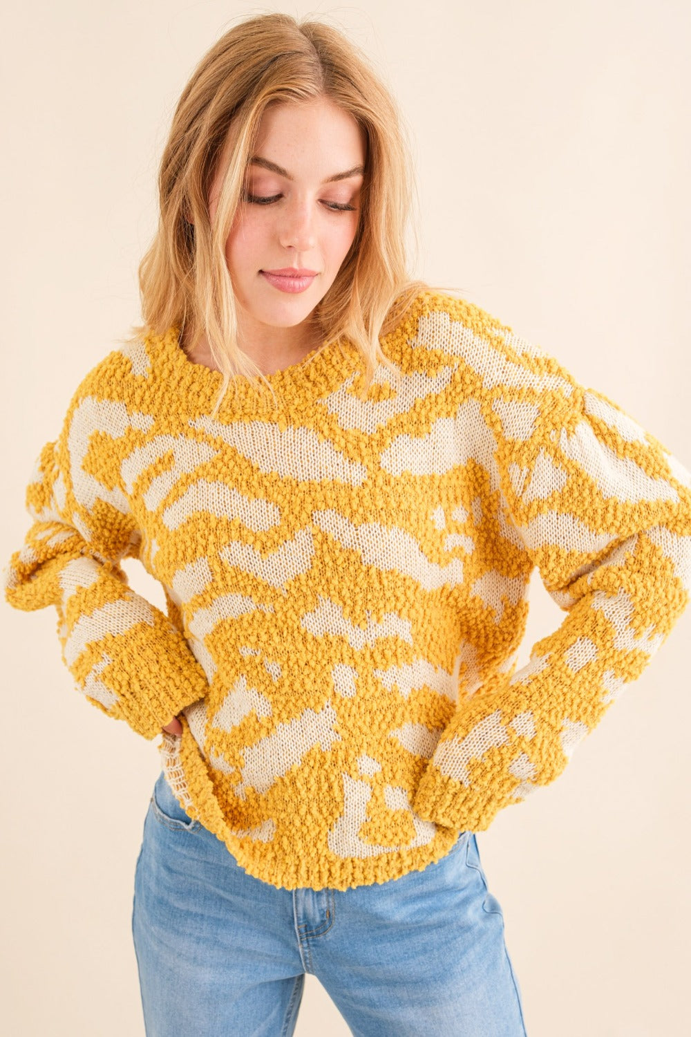 A person with long hair wearing a stylish yellow and white And The Why Full Size Textured Pattern Contrast Sweater and blue jeans stands against a beige background.