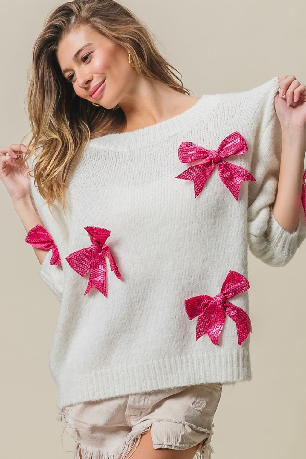 A woman wearing a striking and fashionable BiBi Sequin Bow Puff Sleeve Sweater, paired with light-colored shorts, stands against a plain background.