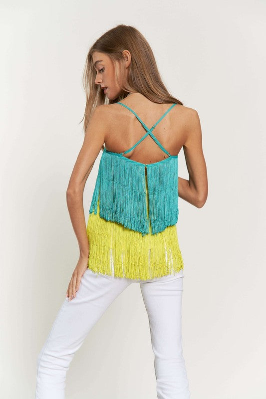 A woman with long brown hair is wearing a Fringe Overlay Cross Straps Party Cami Top featuring blue and yellow layers, paired with white pants, standing against a plain background.