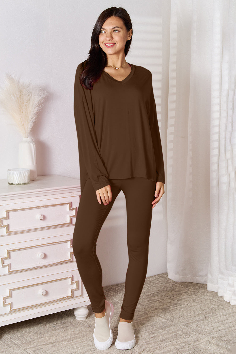 A woman stands indoors, posing in front of a white dresser next to a tall vase with decorative white feathers. She is wearing the Basic Bae Full Size V-Neck Soft Rayon Long Sleeve Top and Pants Lounge Set in bright pink, paired with highly stretchy material and completed with white slip-on shoes.