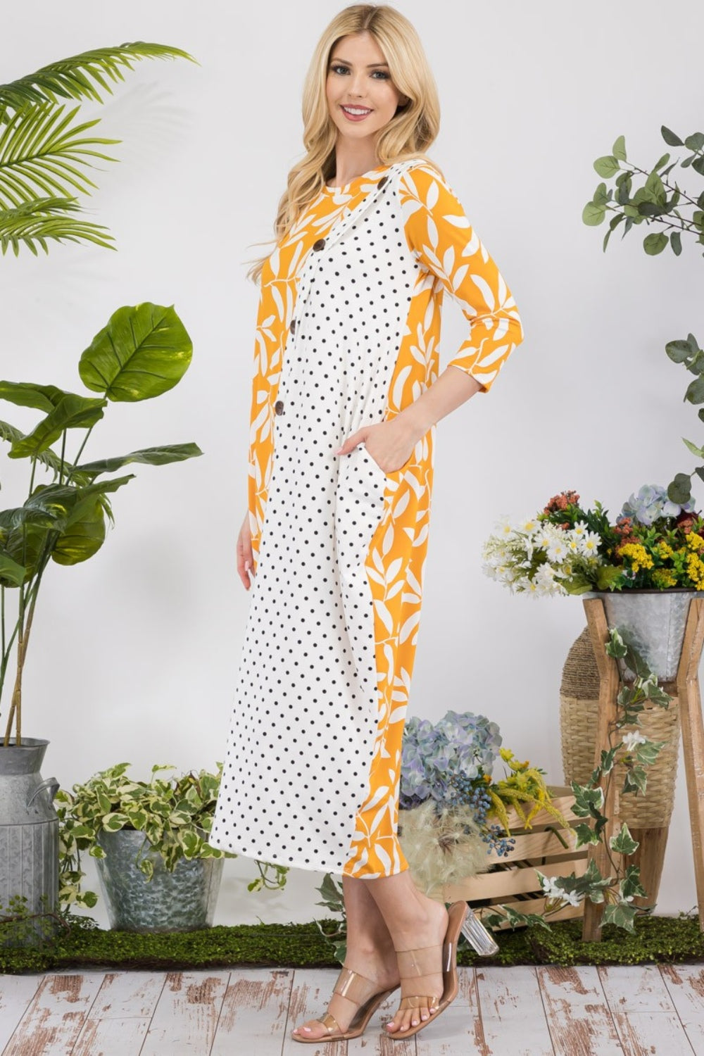 In a garden setting, a woman models the Celeste Full Size Floral Polka Dot Contrast Midi-Dress with Pockets, featuring its versatile yellow design among lush plants and vibrant blooms.