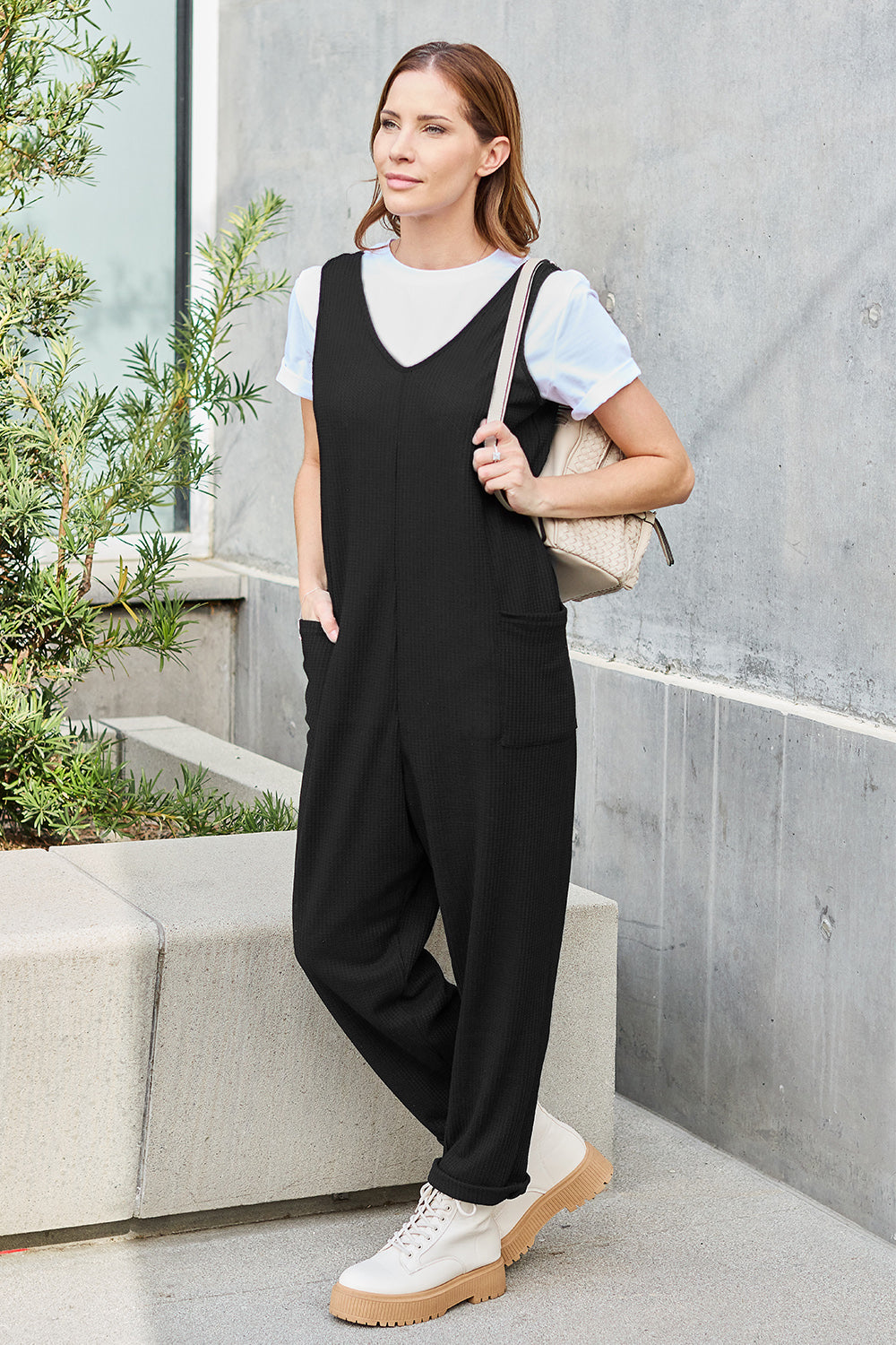 A person stands outdoors wearing the Double Take Full Size Sleeveless Straight Jumpsuit in blue and brown sandals, holding a cream-colored bag. In the background, there's a wooden bench and palm trees.