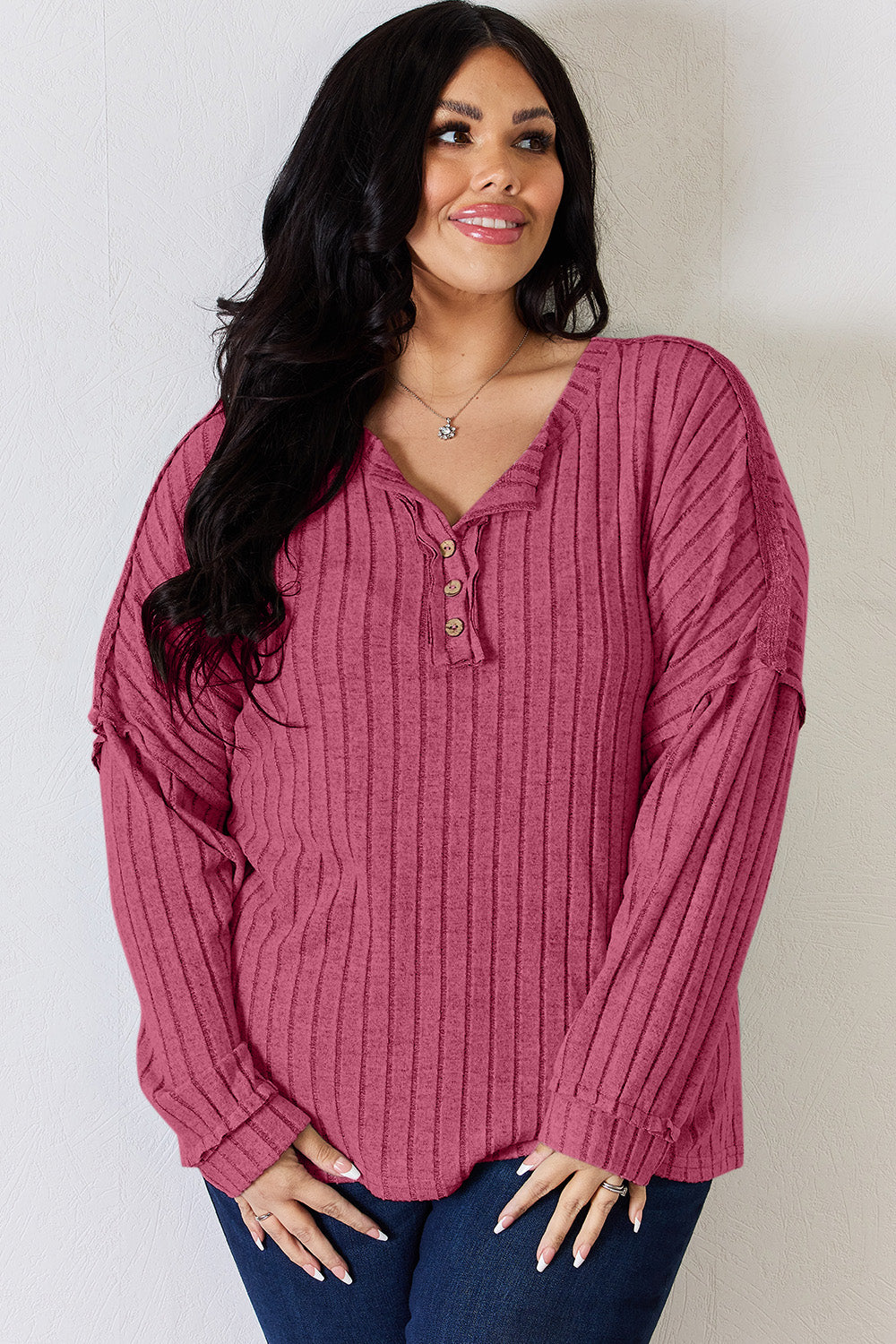 A woman with long, wavy blonde hair wears a slightly stretchy, textured Basic Bae Full Size Ribbed Half Button Long Sleeve T-Shirt in pink, standing against a neutral backdrop.