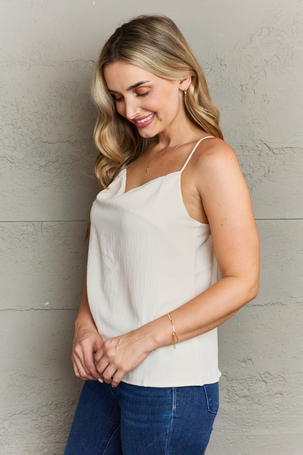 A person with long wavy hair wearing a Ninexis For The Weekend Loose Fit Cami and blue jeans stands against a gray textured wall.