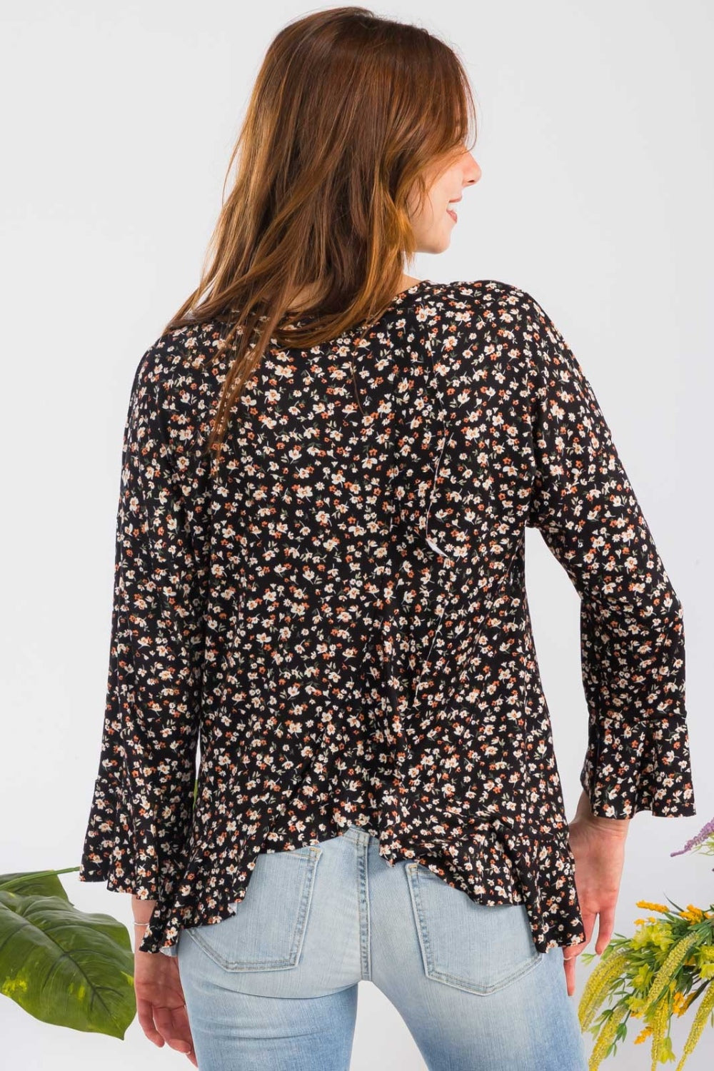 A woman elegantly stands beside colorful flowers against a plain background, embodying feminine fashion in the Celeste Full Size Floral Ruffle Detail Top, which features a round neck and floral patterns, paired with light blue jeans.