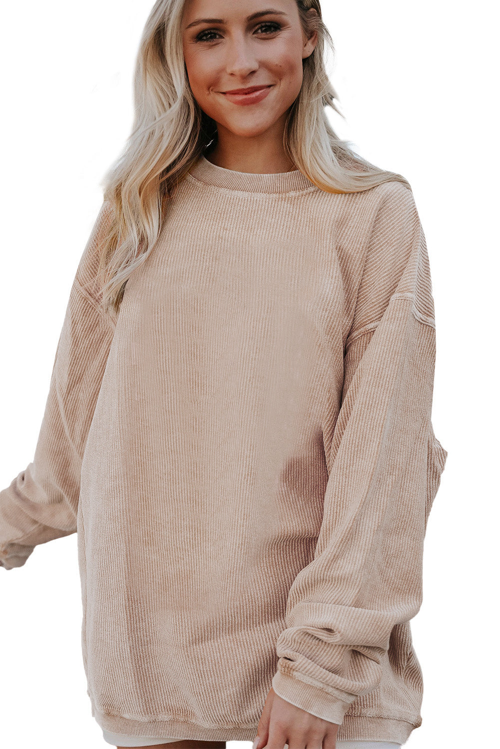 A woman stands outdoors, looking down while touching her hair with one hand. She is wearing an Apricot Ribbed Corduroy Oversized Sweatshirt and white pants, exuding an effortlessly chic and fashionable look.