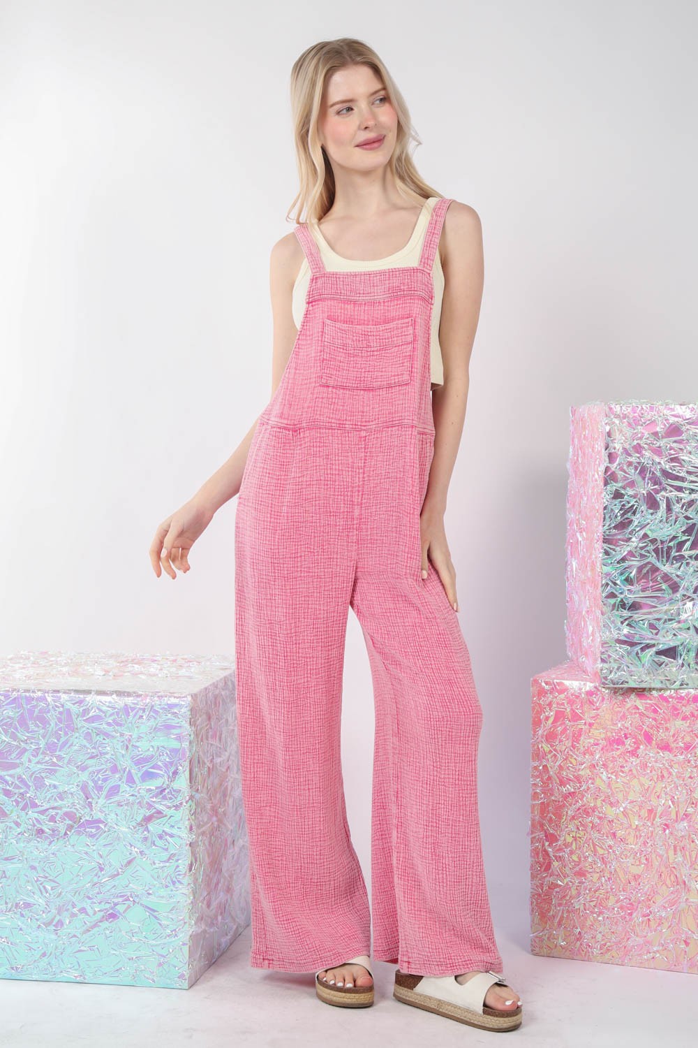 The person is wearing VERY J Texture Washed Wide Leg Overalls in pink with a vintage charm and sandals, standing next to pastel-colored reflective cubes against a white background.