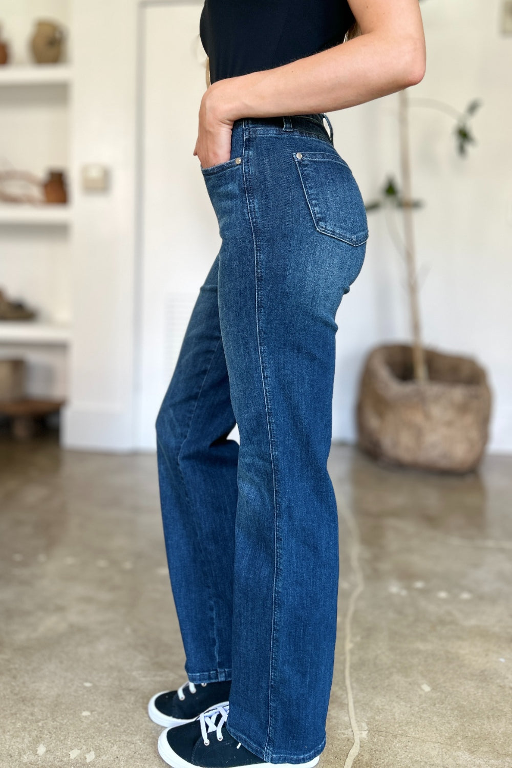 A person wearing a black top and Judy Blue Full Size Tummy Control Straight Jeans stands indoors with one hand on their hip. Their shoes are black with white soles. A plant and shelves are in the background.