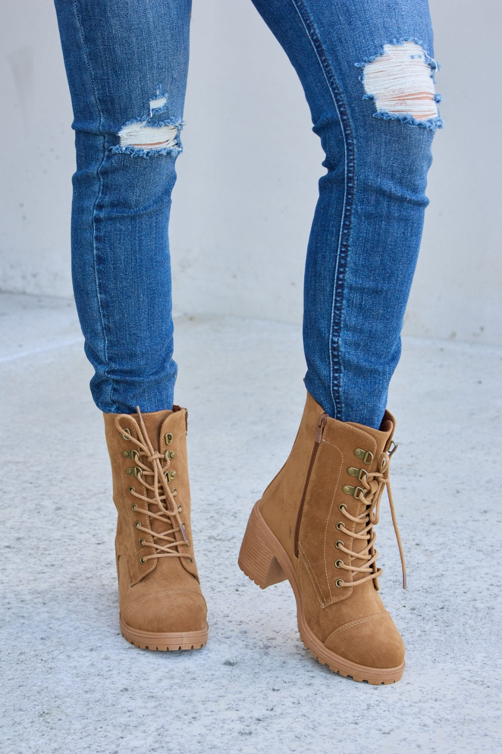 A person wearing distressed blue jeans and Forever Link Lace-Up Zipper Detail Block Heel Boots stands on a concrete surface.