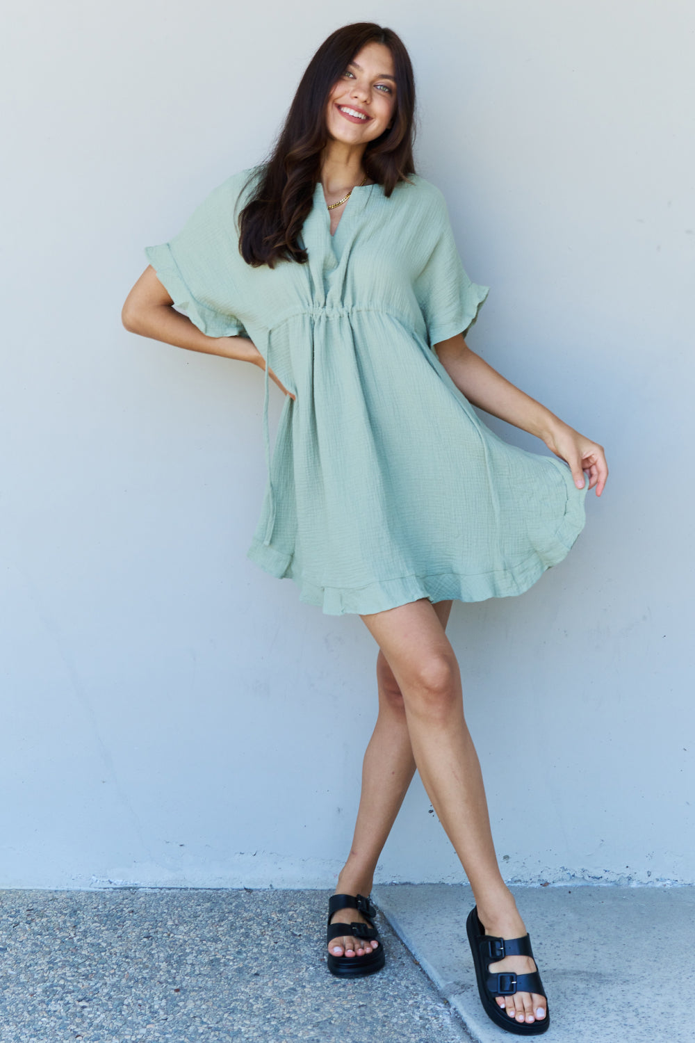 A woman is wearing the Ninexis Out Of Time Full Size Ruffle Hem Dress with Drawstring Waistband in Light Sage. Her long, dark hair falls gracefully as she smiles and looks to her left, standing against a plain background.