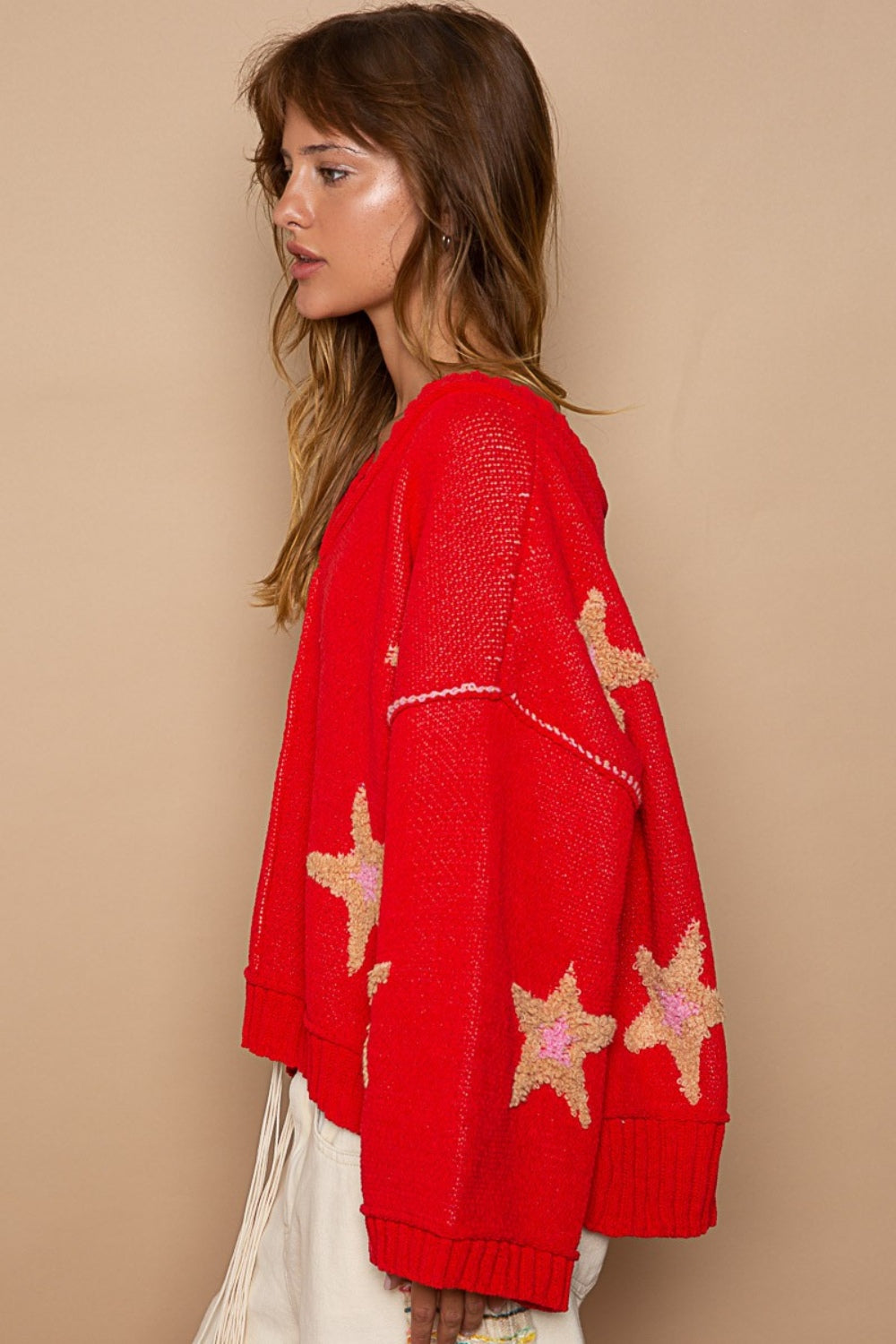 A woman with long hair wears a stylish POL Long Sleeve Star Patch Sweater, paired with light-colored pants, standing against a beige background.