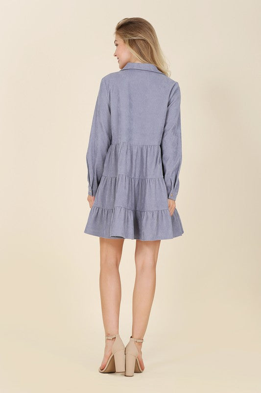 The woman in the Corduroy tiered dress, featuring a deep V-neck, smiles as she gently touches her forehead, showcasing the light blue fabric cascading elegantly.