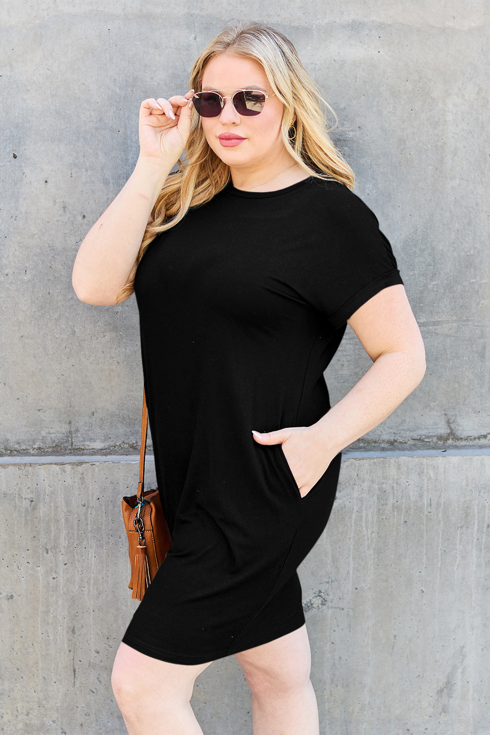 A woman wearing a Basic Bae Bamboo Full Size Round Neck Short Sleeve Dress with Pockets and sunglasses holds the strap of a light-colored backpack while standing against a concrete wall.