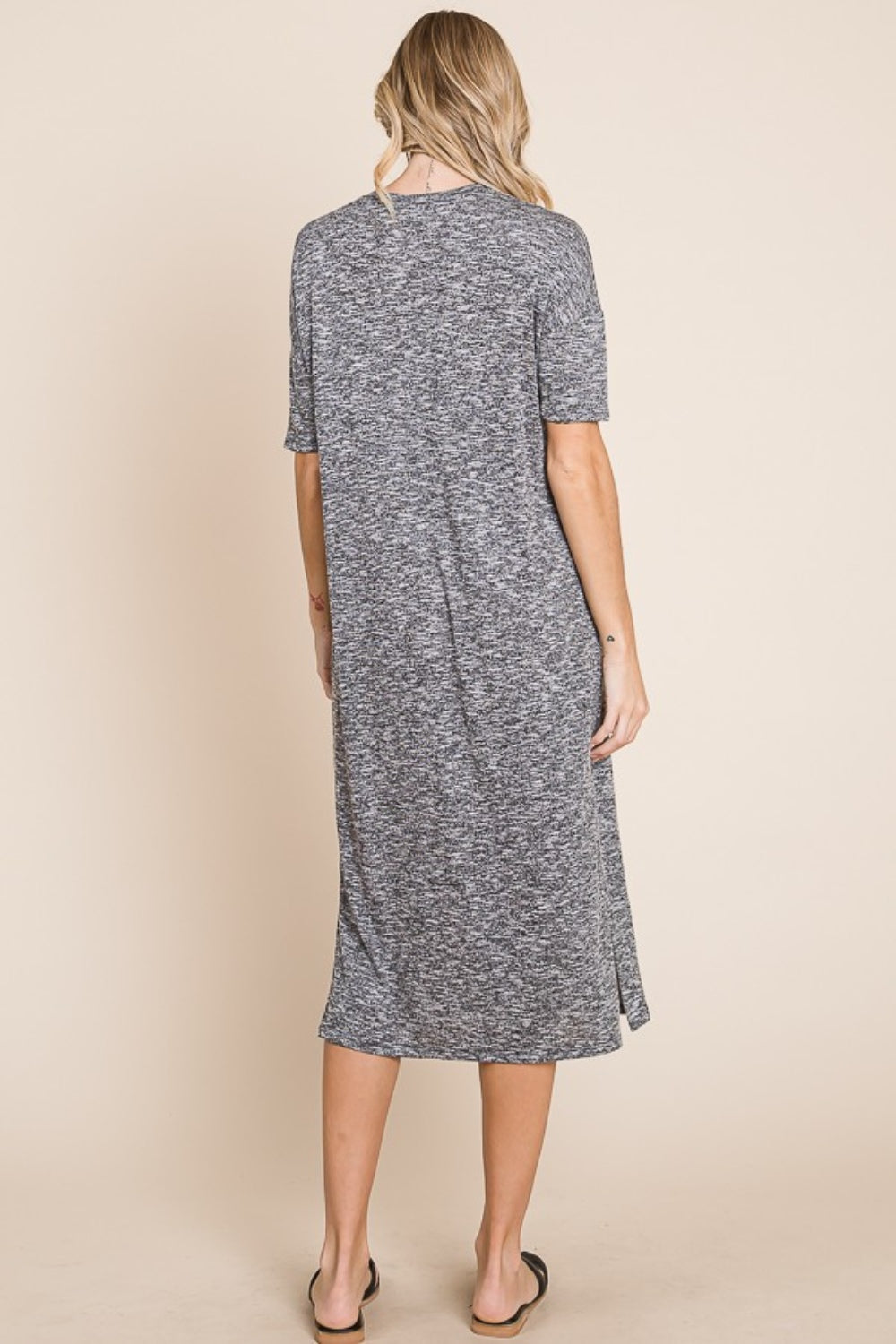 A woman wearing the BOMBOM Slit Round Neck Half Sleeve Midi Dress, a knee-length, grey midi dress featuring short sleeves and a slit detail at the hem, paired with black slip-on sandals, stands in front of a neutral background.