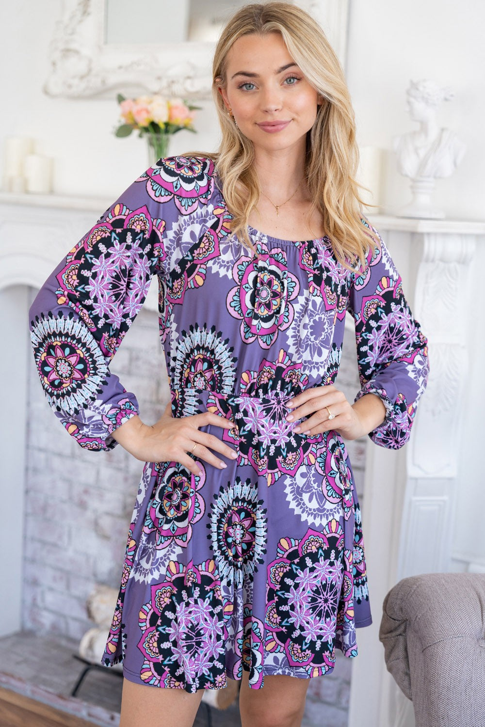 A woman stands indoors wearing the stylish White Birch Printed Long Sleeve Mini Dress with Short Liner, featuring a vibrant floral pattern. She poses with her hands on her hips, smiling at the camera.