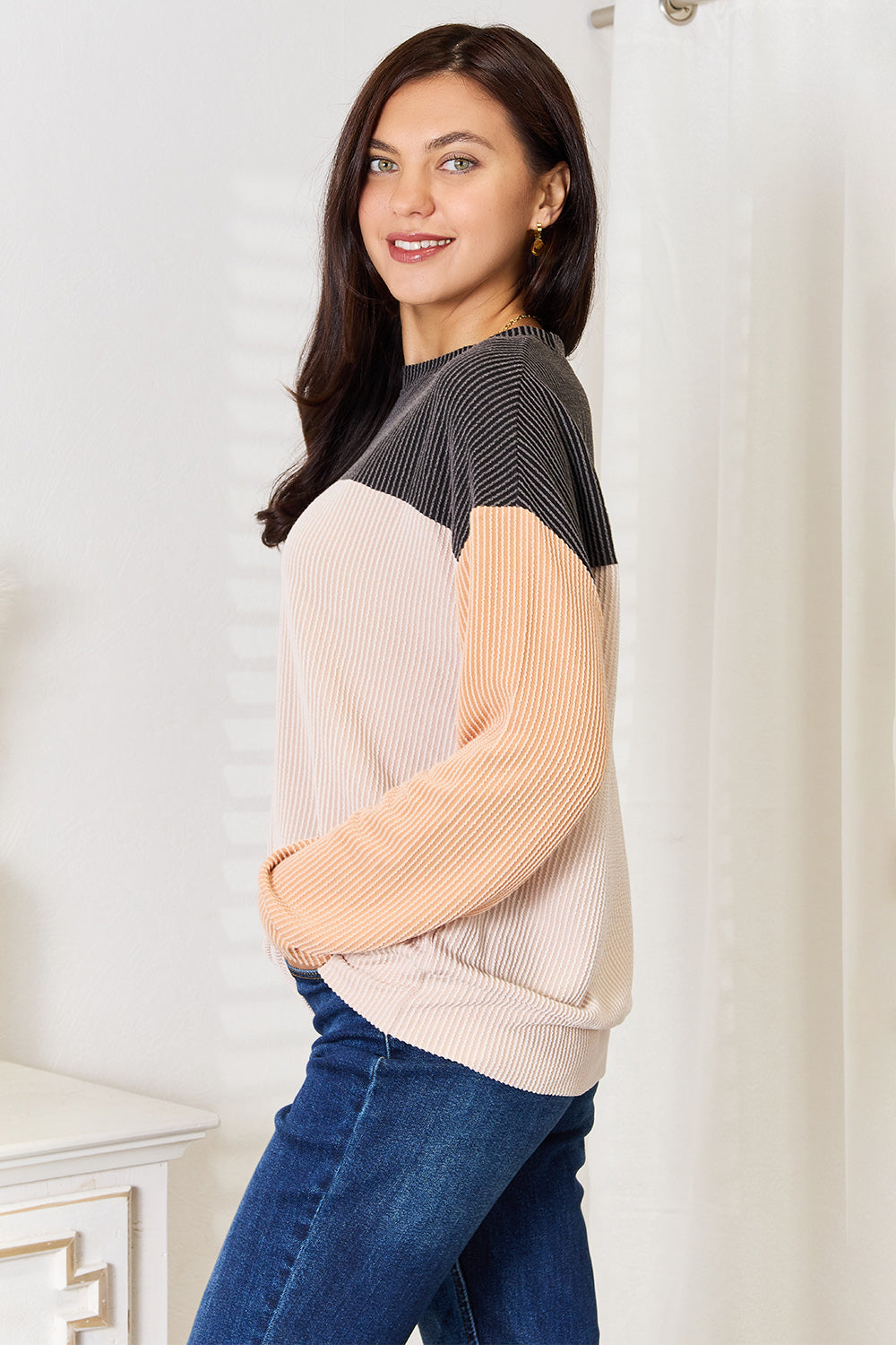 A woman with long brown hair stands indoors, smiling, and wearing a Double Take Color Block Dropped Shoulder T-Shirt paired with jeans. She is positioned near a small table with a white vase containing dried pampas grass, epitomizing the perfect casual wardrobe.